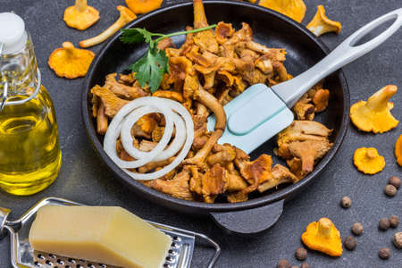 Fried chanterelle mushrooms, spatula and chopped onion in pan. Raw chanterelles, oil in bottle and cheese on table. Black background.の写真素材