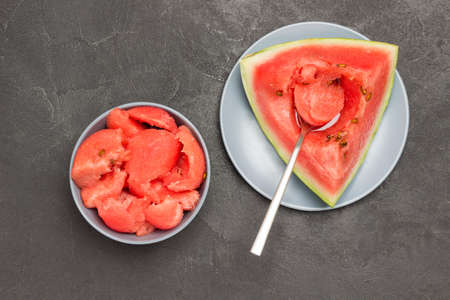 Spoon in pulp of ripe watermelon. Pieces of watermelon in gray bowl. Flat lay. Black backgroundの写真素材