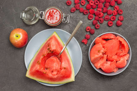 Spoon in pulp of ripe watermelon. Pieces of watermelon in gray bowl. Watermelon juice in glass jar. Apple and raspberry on table. Flat lay. Black backgroundの写真素材