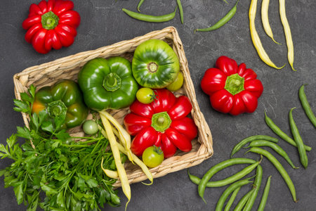 Pepper, green tomato and bunch of parsley in wicker basket. Red peppers and green beans on table....