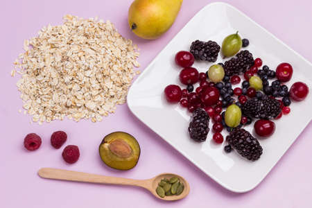 Oat flakes, half plum, pear and pumpkin seeds in spoon on table. Plate with frozen berries. Flat lay. Pink backgroundの写真素材
