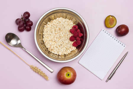 Oatmeal and raspberries in metal bowl. Notepad and pen. Grapes, plum and apple on table. Flat lay. Pink backgroundの写真素材