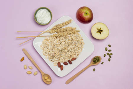 Spikelets of wheat and oat flakes in plate. Wooden spoons, pumpkin seeds and nuts on the table. Two halves of an apple. Flat lay. Pink backgroundの写真素材