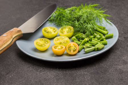 Chopped celery stalks, green tomatoes and dill on gray plate. Kitchen knife. Black background. Top viewの写真素材