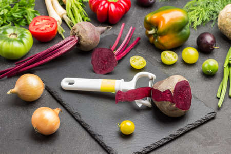Vegetable peeler and beets on cutting board. Tomatoes, peppers and greens on table. Black background. Top viewの写真素材