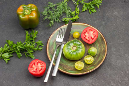 Knife and fork, red and green tomato on plate. Pepper and sprig of parsley on table. Black background. Top viewの写真素材