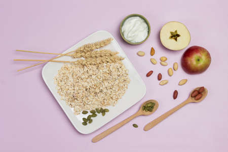 Spikelets of wheat and oat flakes in plate. Wooden spoons, pumpkin seeds and nuts on table. Two halves of apple. Flat lay. Pink backgroundの写真素材