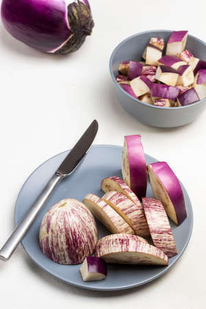 Sliced eggplant and knife on gray plate. Whole purple eggplant with dry leaves on table. Top view. White background.の写真素材