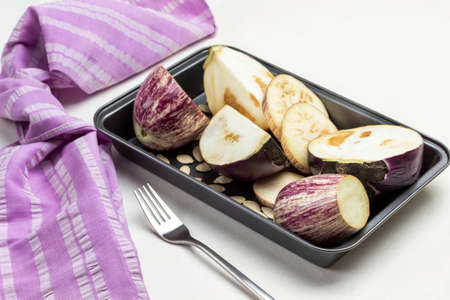 Eggplant sliced into wedges in tray.  Napkin and fork on table. Top view. White background.の写真素材