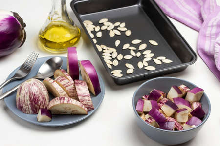 Sliced eggplant in gray bowl. Pumpkin seeds on pallet. Oil in bottle. Top view. White background.の写真素材
