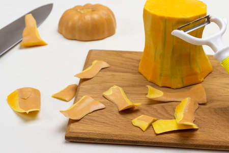 Peeled pumpkin, peeling waste and vegetable peeler on cutting board. White background. Close upの写真素材