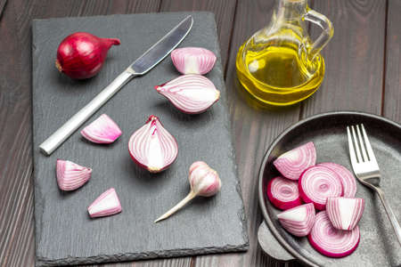 Chopped onion and knife on cutting board. Sliced purple onions and fork in frying pan. Bottle of olive oil. Dark wooden background. Top viewの写真素材