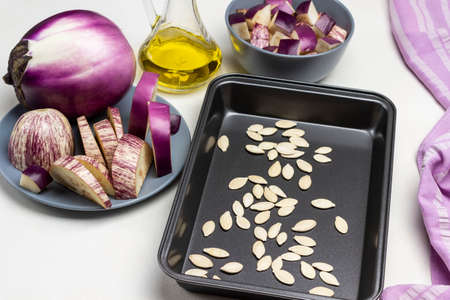 Pumpkin seeds on pallet. Oil in bottle. Sliced eggplant in gray bowl. Whole purple eggplant on plate. Top view. White background.の写真素材