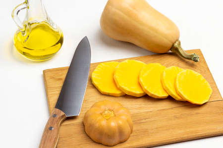Sliced pumpkin and knife on cutting board. Whole pumpkin and bottle of oil on table. White background. Top view.の写真素材