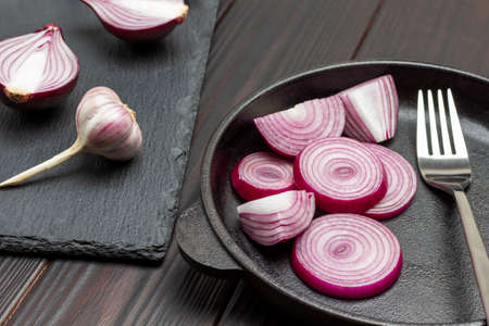 Sliced purple onions into rings in frying pan. Onion halves and head of garlic on cutting board. Dark wooden background. Top viewの写真素材
