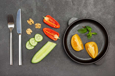 Chopped yellow tomatoes in frying pan. Cucumber and slices of red pepper, fork and knife on table. Black background. Flat lay.の写真素材