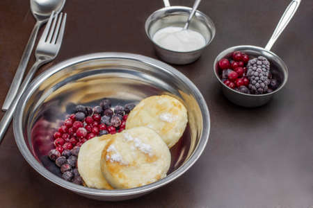 Cottage cheese pancakes and berries in metal bowl. Frozen berries and yoghurt in small metal bowl. Fork and spoon. Rusty metal background. Top view.の写真素材