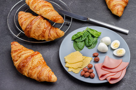 Ham, cheese, nuts and spinach on gray plate. Fresh croissants and knife. Top view. Black background.の写真素材