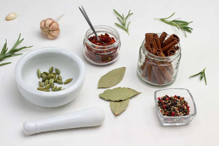 Cinnamon sticks in jar. Cardamom in mortar and pestle on table. Allspice in glass bowl. Paprika petals in jar. Bay leaf and rosemary on table. Top view. White background.の写真素材