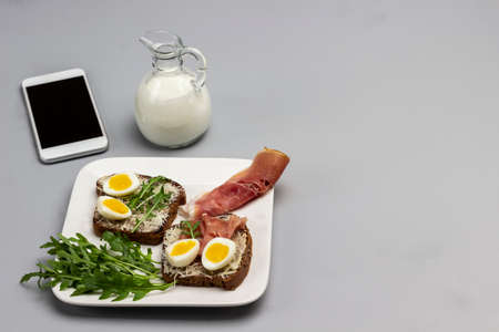 Boiled egg and grated cheese sandwich, fork on white plate. Creamer with cream. Mobile on table. Copy space. Top view. Gray background.の写真素材