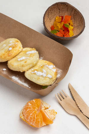 Cottage cheese pancakes in disposable plate. Mandarin, Candied fruits in coconut shells, fork and knife on table. White background. Top viewの写真素材