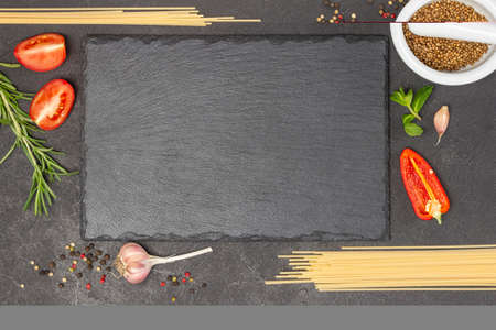 Black stone cutting board on black background. Coriander seeds in mortar with pestle. Pasta, halves of tomato, garlic and rosemary. top viewの写真素材