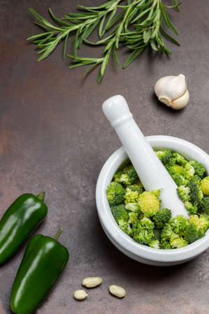Broccoli in mortar and pestle. Two green peppers and sprigs of rosemary on rusty metal background. top viewの写真素材