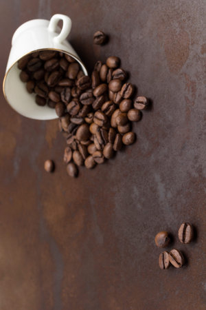 Roasted coffee beans are scattered from black cup onto table. Rusty metal background. flat lay. copy...