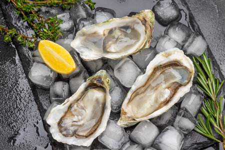 Opened oyster shells with ice on black background. Lemon and greens on table. flat lay. black...