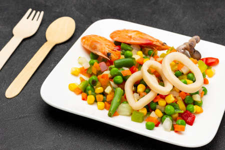 Shrimp and squid rings with mixed vegetables on white plate. Spoon and fork on table. top view. black backgroundの写真素材