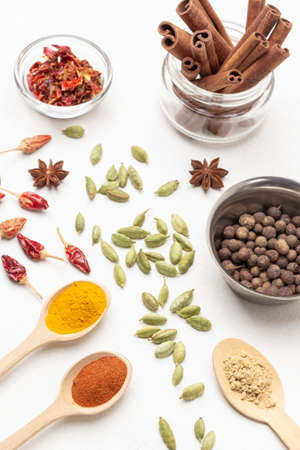 Allspice in metal bowl and cinnamon sticks in jar. Ground paprika, turmeric and ginger spices in wooden spoons. Cardamom seeds and dry pepper pods on table. top view. white backgroundの写真素材