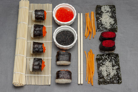 Nori sushi with caviar on bamboo mat and on table. Black sesame and red caviar in bowls. Chopsticks. flat lay. dark backgroundの写真素材