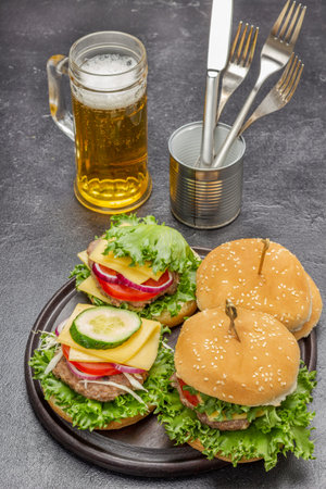 Cheeseburger with greens on round board. Glass mug with beer. Knife and fork in tin. top view. black backgroundの写真素材