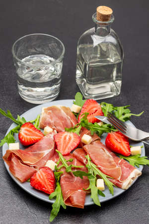 Spanish jamon with cheese, strawberries and arugula on gray plate. Fork and knife on plate. Glass bottle and glass of water. top view. black backgroundの写真素材