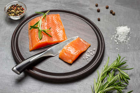 Pieces of salmon and kitchen knife on cutting board. Rosemary, salt and allspice on table. top view. brown backgroundの写真素材