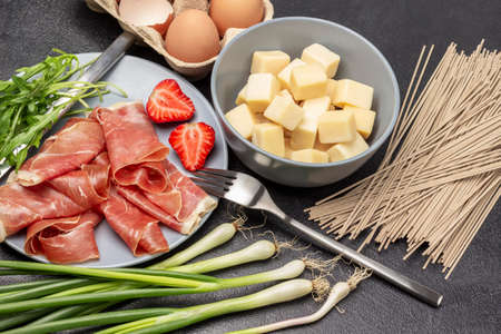 Spanish jamon with strawberries and arugula on gray plate. Eggs in a box. Noodles, green onions on table. Cheese and mustard in bowls. top view. black backgroundの写真素材