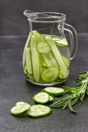 Cucumber infused water in glass jug. Sprig of rosemary on table. Black background.の写真素材