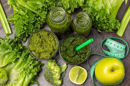 Green smoothie in glasses and bottles. Measuring tape around an apple. Lettuce leaves and celery stalks on the table. flat lay. gray backgroundの写真素材