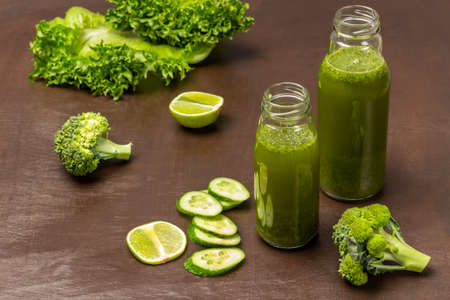 Two green smoothie bottles. Slices of cucumber, lime, broccoli and lettuce on a brown background. top viewの写真素材
