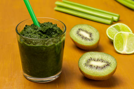 Green smoothie and straw in glass. Kiwi and lime slices, celery stalks on table. Bright mustard background. top viewの写真素材