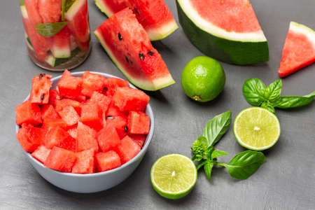 Sliced watermelon pulp in bowl. Watermelon slices, lime halves and sprig of basil on table. top view. gray background.の写真素材