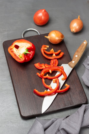 Chopped red pepper, kitchen knife and half pepper on cutting board. Onion, tomato and gray napkin on table. top view. gray background.の写真素材