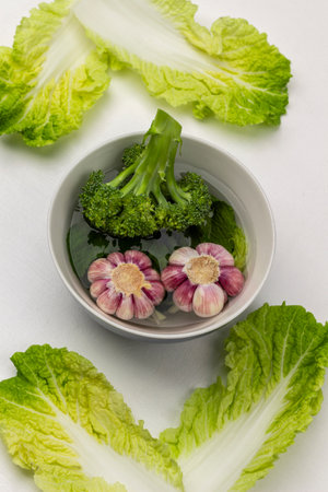 Two heads of garlic and broccoli in a bowl of water. Yellow-green lettuce leaves. flat lay. white background.の写真素材