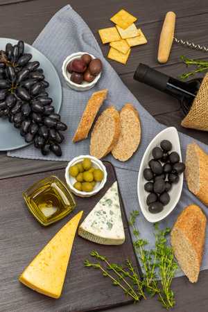 Cheese, olive oil and olives on a chalkboard. Black olives, a bunch of black grapes and a sliced baguette on a gray napkin. a bottle of wine. flat lay. Dark wooden background.の写真素材