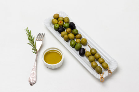 Green olives on skewers. Olives on white plate. Olive oil in bowl. Fork on table. white background.の写真素材
