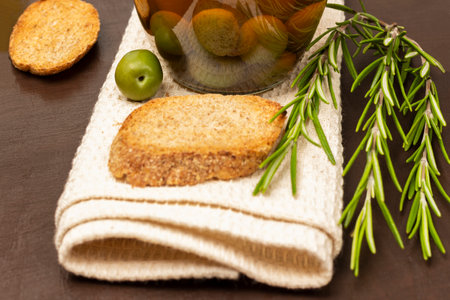 Green olive and a piece of baguette on a napkin. Canned olives in a jar. Rosemary sprigs. brown backgroundの写真素材