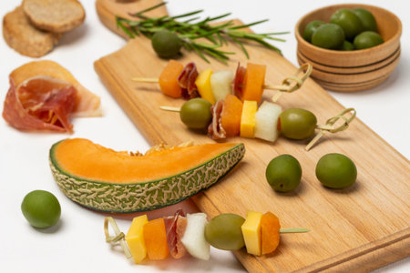 A slice of melon. Cheese, olives, jamon on a skewer. Green olives in a wooden bowl. top view. white background.の写真素材