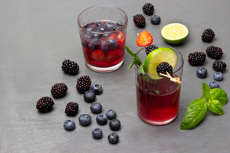 Blackberries, blueberries, green basil leaves on table. Two glasses of berry drink. Blackberry and lime wedge on skewer. top view. gray background.の写真素材