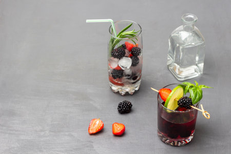 Ice, rosemary and berries in glass. Infused summer drink with blackberries and green basil in glass. water bottle. Strawberries and blackberries on table. copyspace. gray backgroundの写真素材