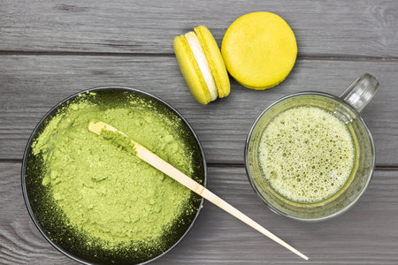 Two macaroni cakes and mug of matcha green tea. Measuring spoon in bowl with matcha powder. Dark wooden background.の写真素材